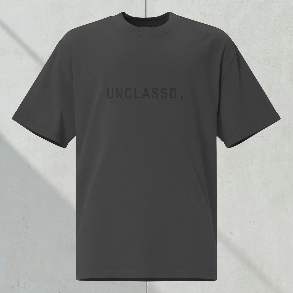 UNCLASSD CORE Oversized Tee in faded black with tonal “UNCLASSD.” chest print, ghost mockup against dark concrete background.